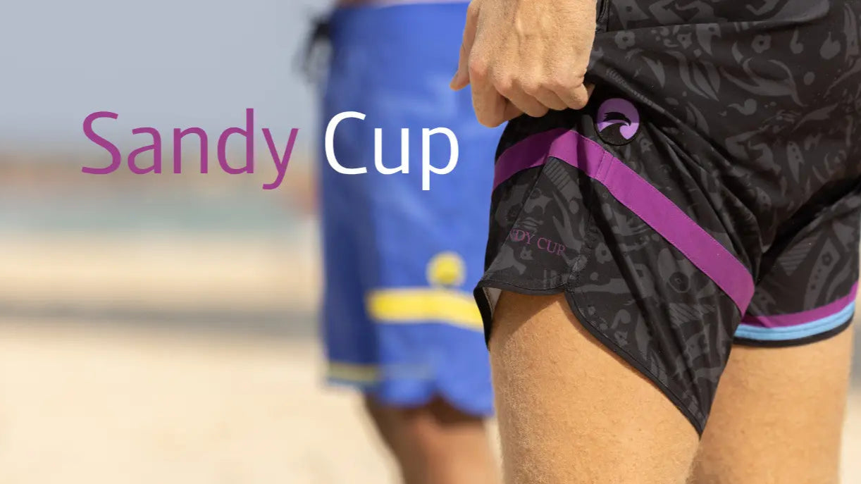 Person wearing black patterned shorts with a purple waistband on a beach, with 'Sandy Cup' text.